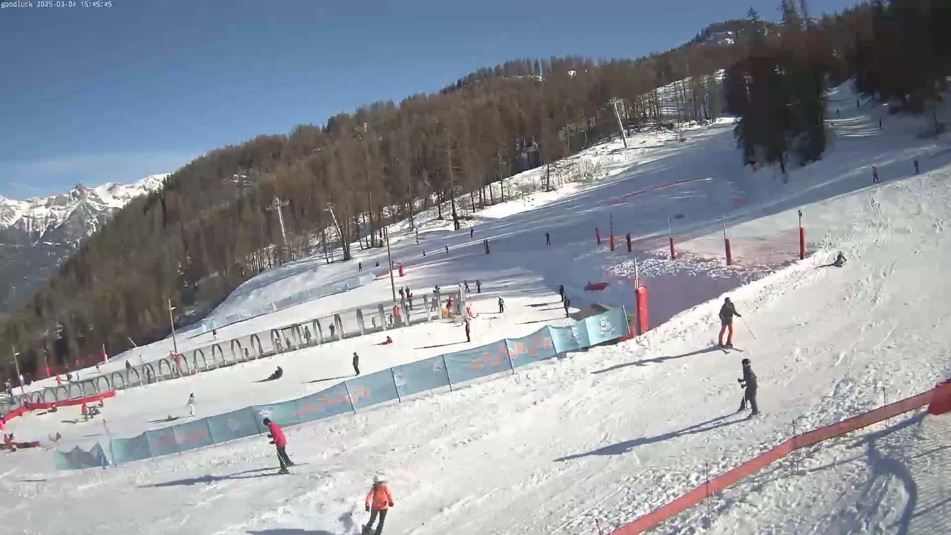 A sunny day on a snow-covered ski slope with several skiers and snowboarders,  surrounded by trees and mountains in the background.