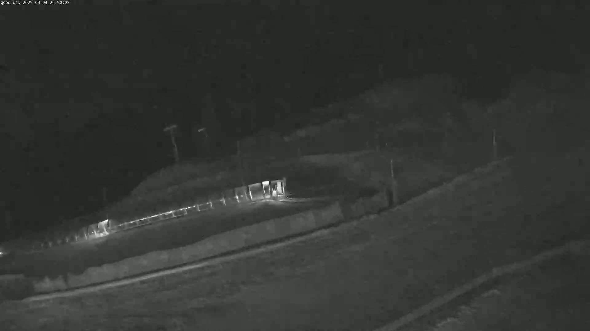 A nighttime, low-light image shows a person walking toward a lit structure near a fence, in what appears to be a parking lot or similar area.