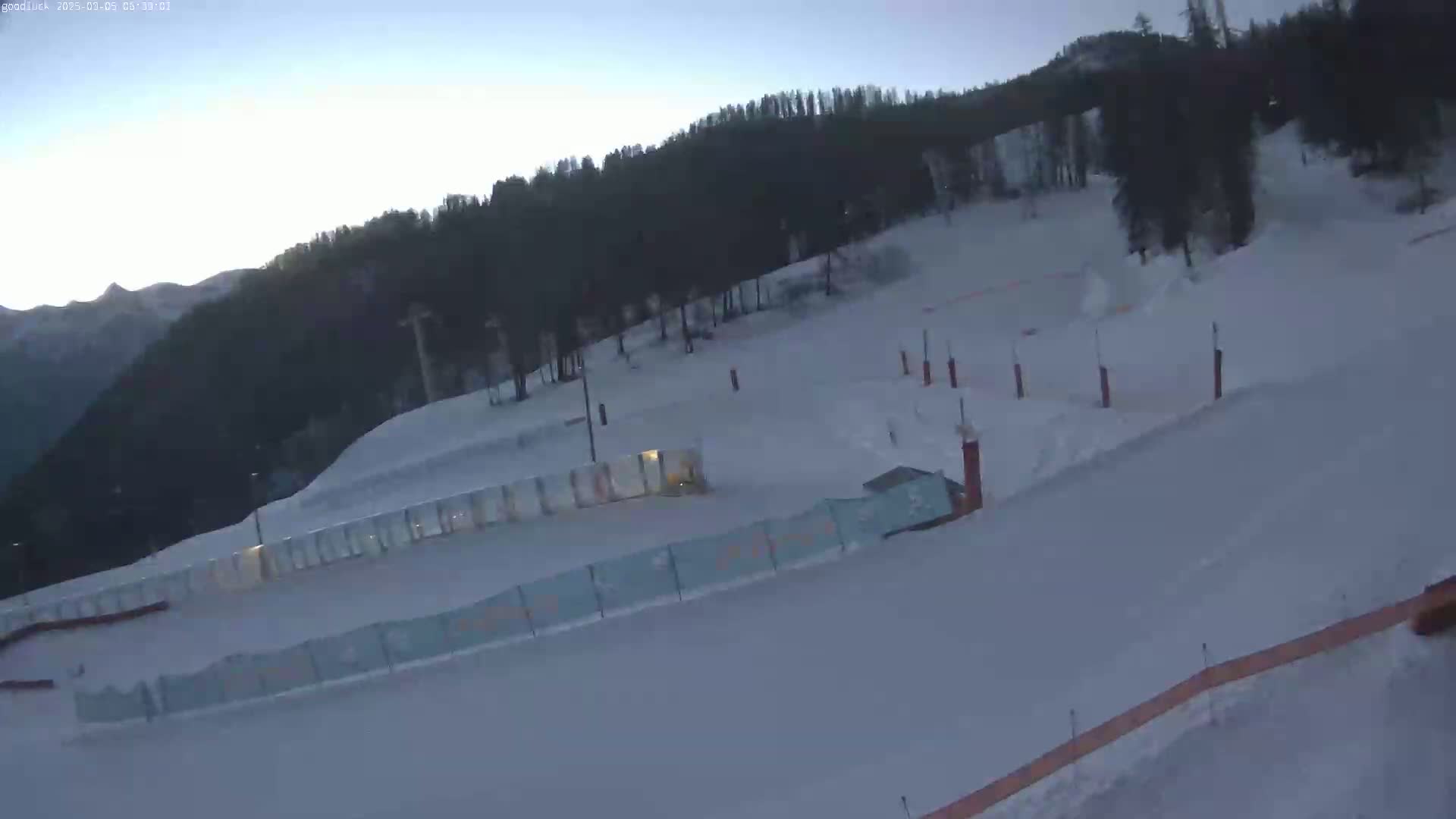 A snow-covered ski slope with a forested mountain backdrop under a clear, twilight sky.