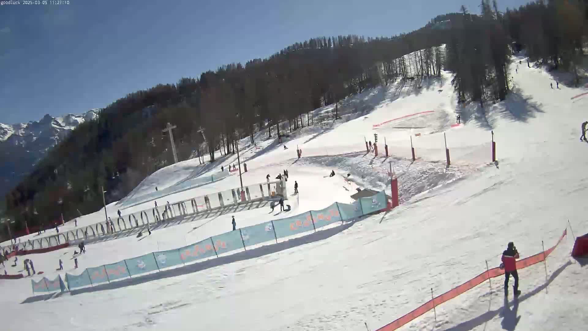 A sunny day at a snow-covered ski slope with several skiers and snowboarders visible, mountains in the background.
