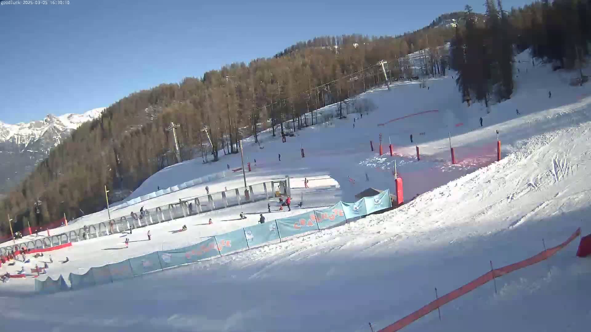 A snow-covered ski slope with several people skiing and snowboarding, under a clear sunny sky, is surrounded by trees and mountains in the background.