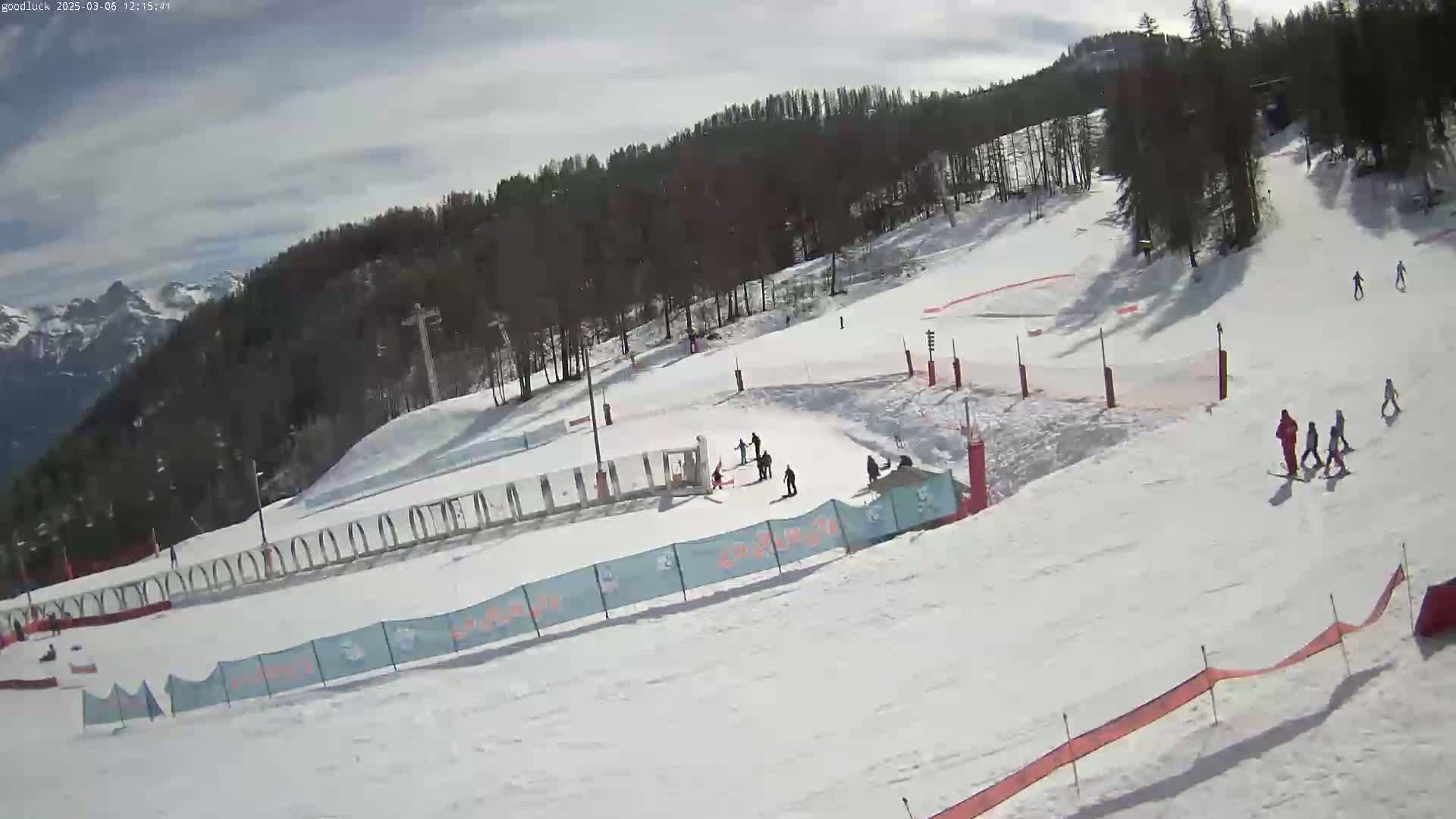 A snow-covered ski slope with several skiers and snowboarders, under a partly cloudy sky, is surrounded by trees and mountains in the background.