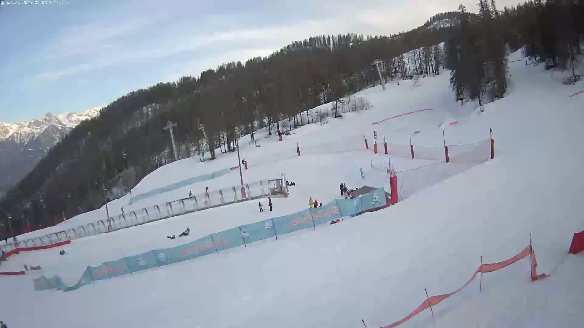 A snow-covered ski slope with several people skiing and snowboarding, under a mostly clear sky.