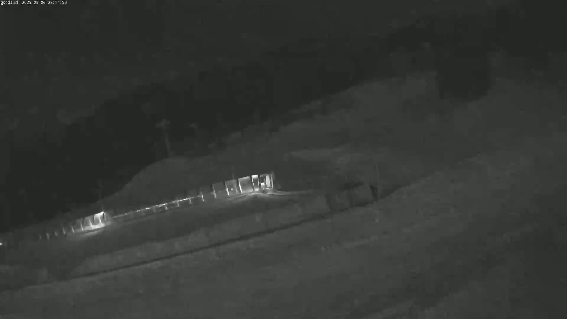 A nighttime, low-light image shows a paved area with a fence and a structure, possibly a building or gate, in the middle distance.