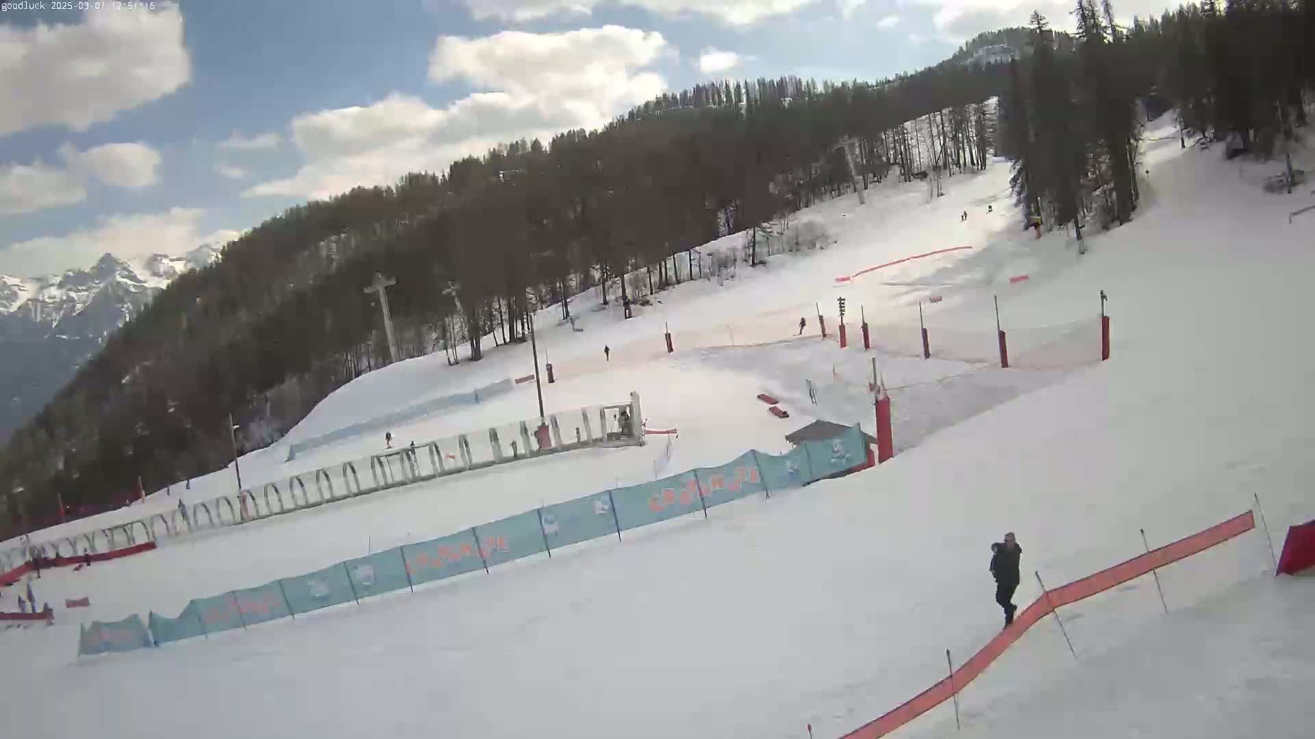 A snow-covered ski slope with several skiers, a lift, and trees in the background under a partly cloudy sky.