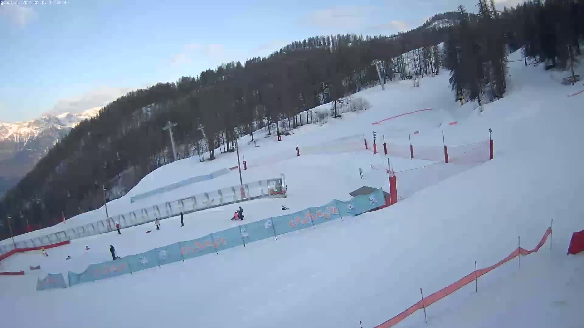 A snowy ski slope with several people skiing and snowboarding, surrounded by trees and mountains under a clear sky.