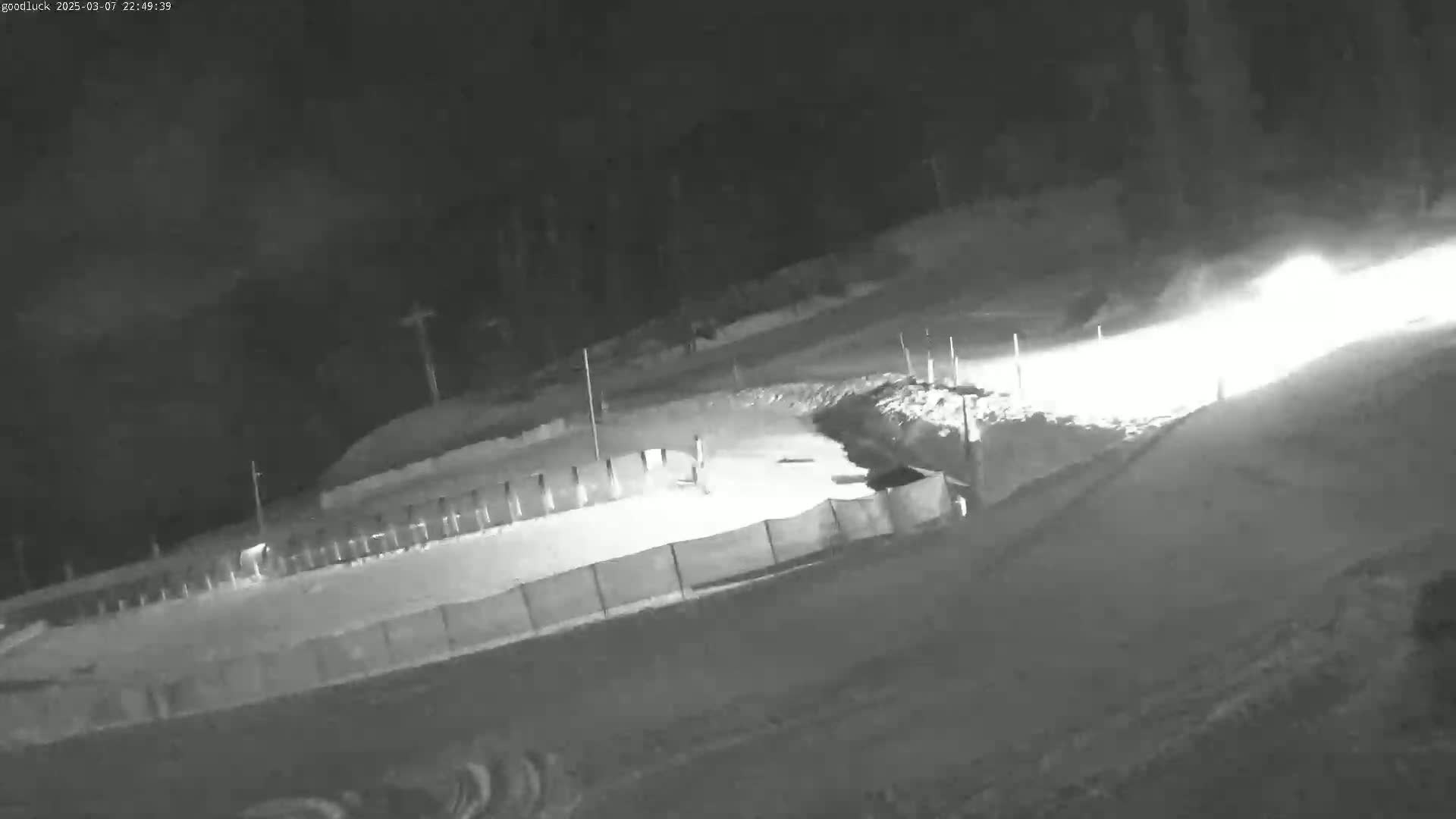 A nighttime, snowy scene shows a fenced-in area with bright headlights approaching from the upper right.