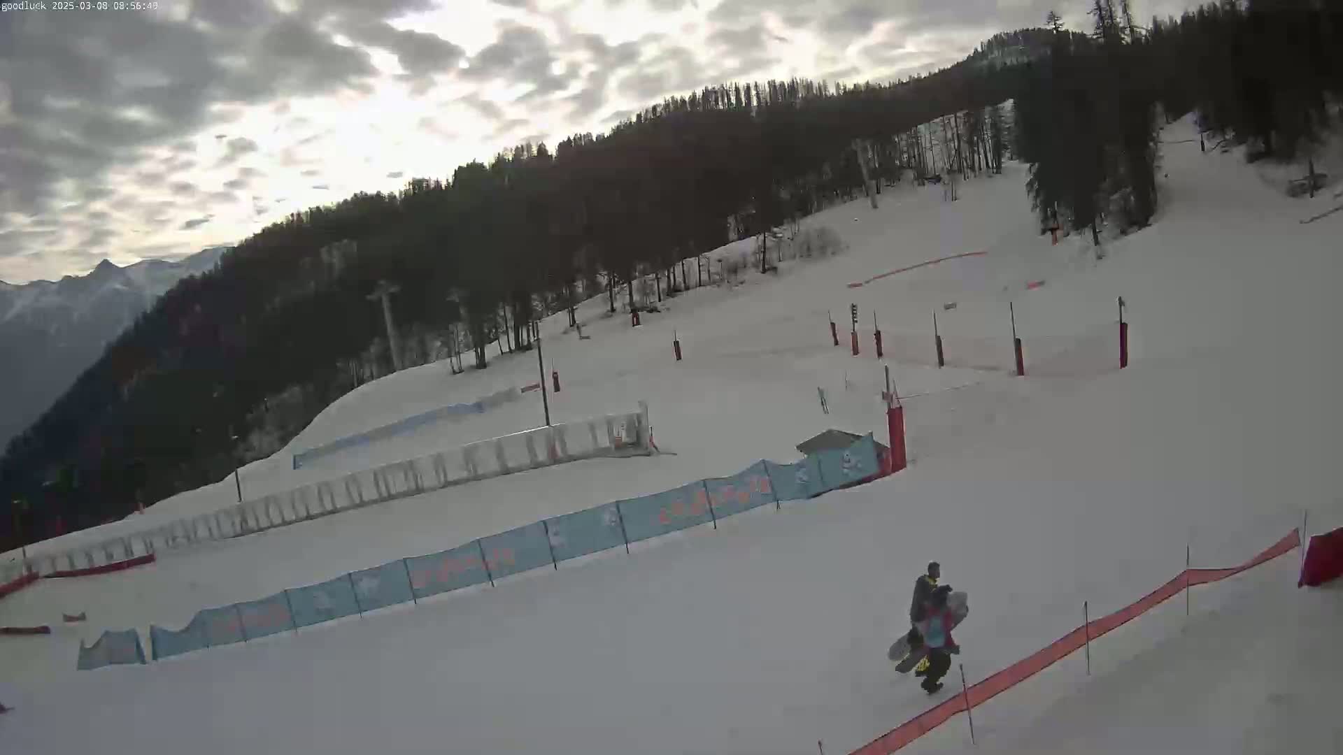 A person carries a snowboard down a snow-covered ski slope under an overcast sky, with a forested mountain in the background.