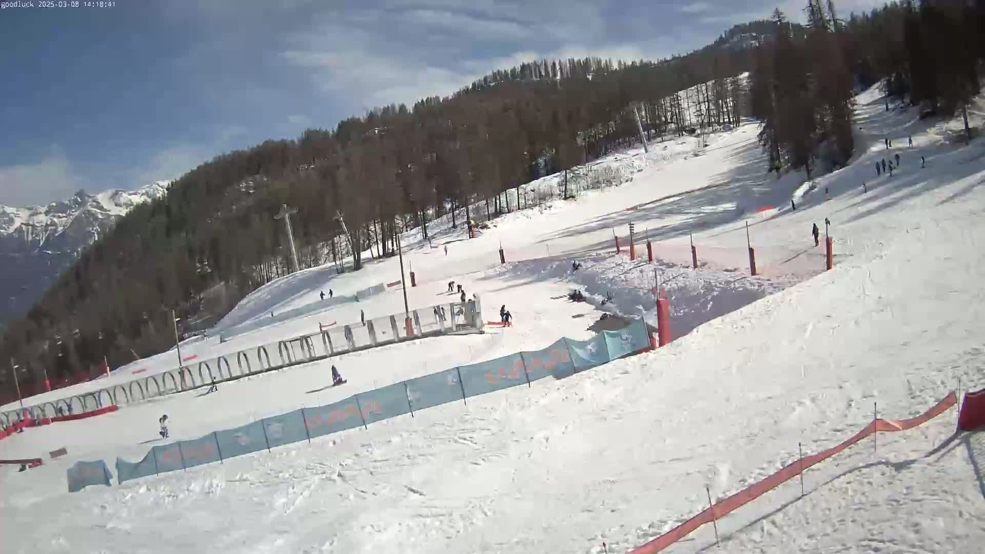 A snowy ski slope with several skiers and snowboarders, under a mostly sunny sky, is situated against a backdrop of snow-covered mountains and trees.