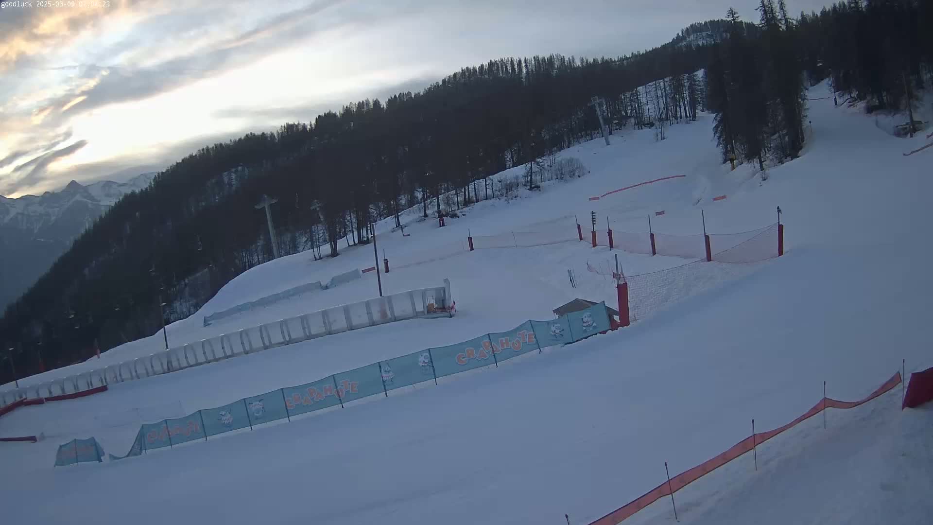A snow-covered ski slope with trees and mountains in the background under a cloudy sky.