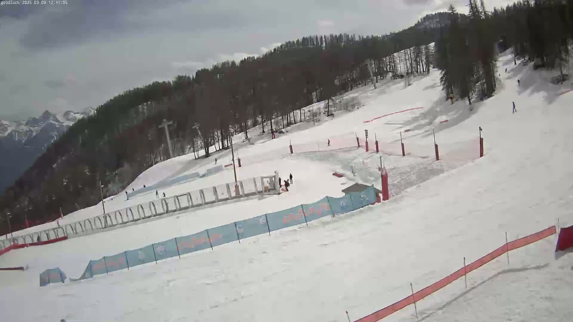 A snow-covered ski slope with several skiers and snowboarders, a ski lift in the background, and a mostly cloudy sky.