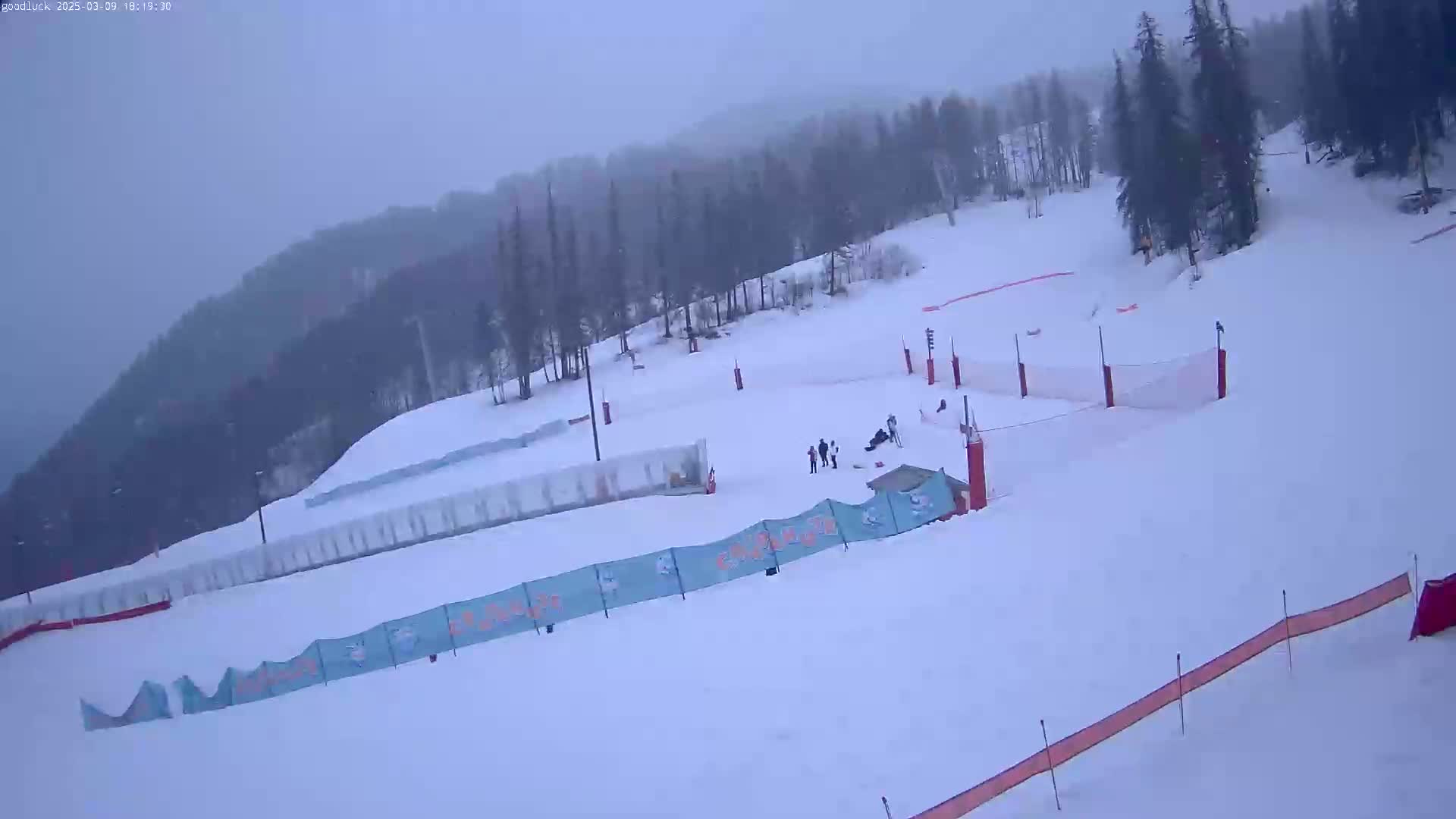 A snow-covered ski slope with several people near the bottom, under an overcast sky.
