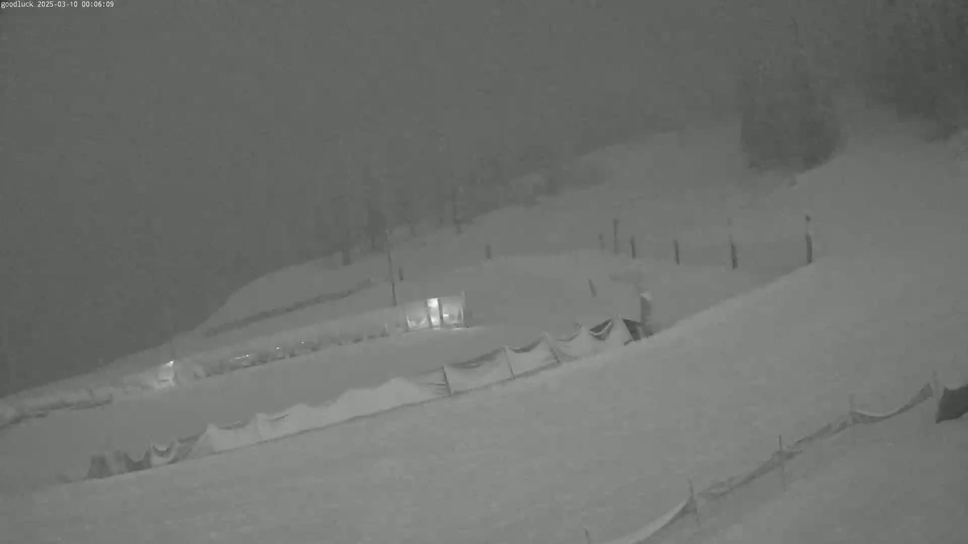 A snow-covered landscape at night is visible under heavy snowfall, with a dimly lit structure and snow fences in the mid-ground.