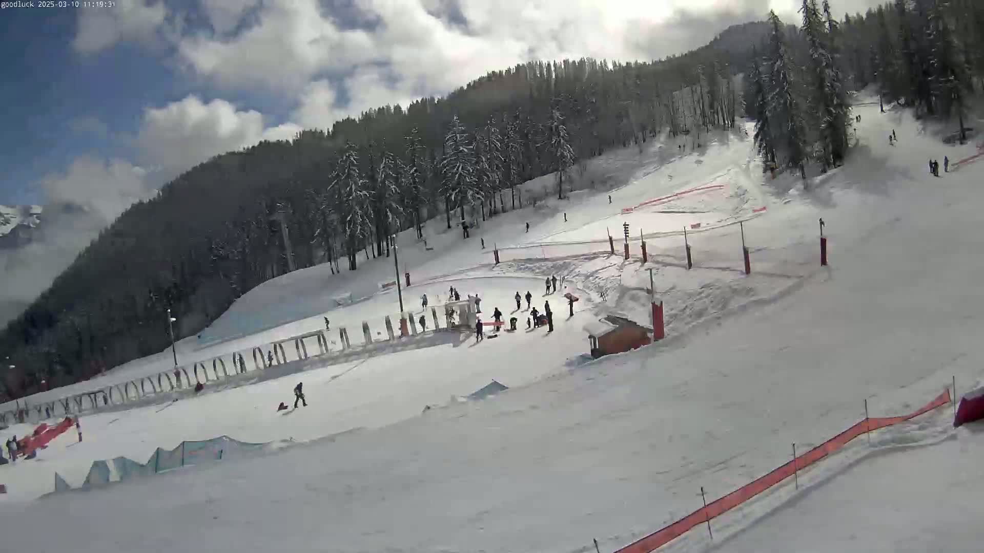 A snow-covered ski slope with several skiers and snowboarders, under a partly cloudy sky.