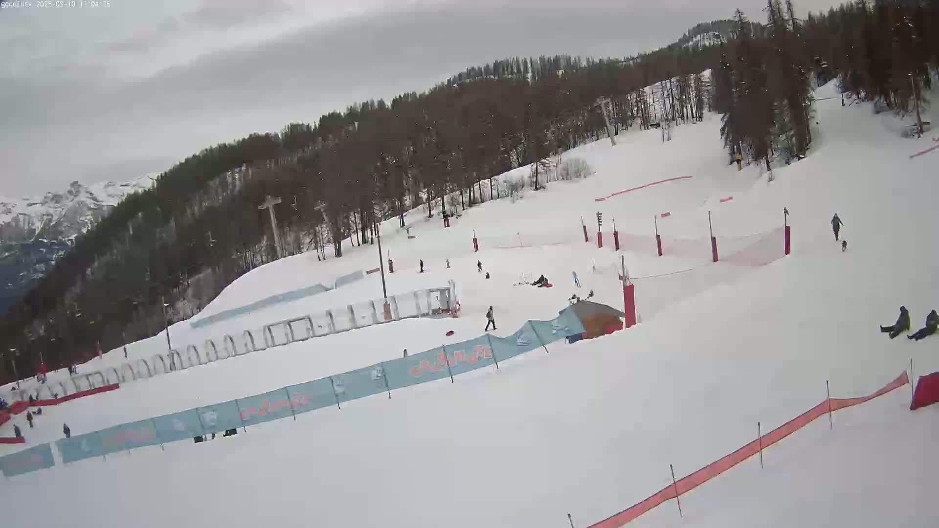 A snow-covered ski slope with several people skiing, snowboarding, and walking, under an overcast sky.