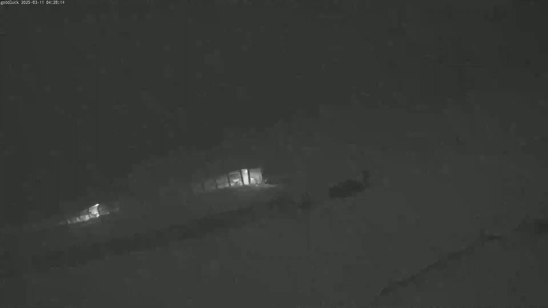 A dimly lit, snowy nighttime scene shows a few structures with faint interior lights visible through the heavy snowfall.