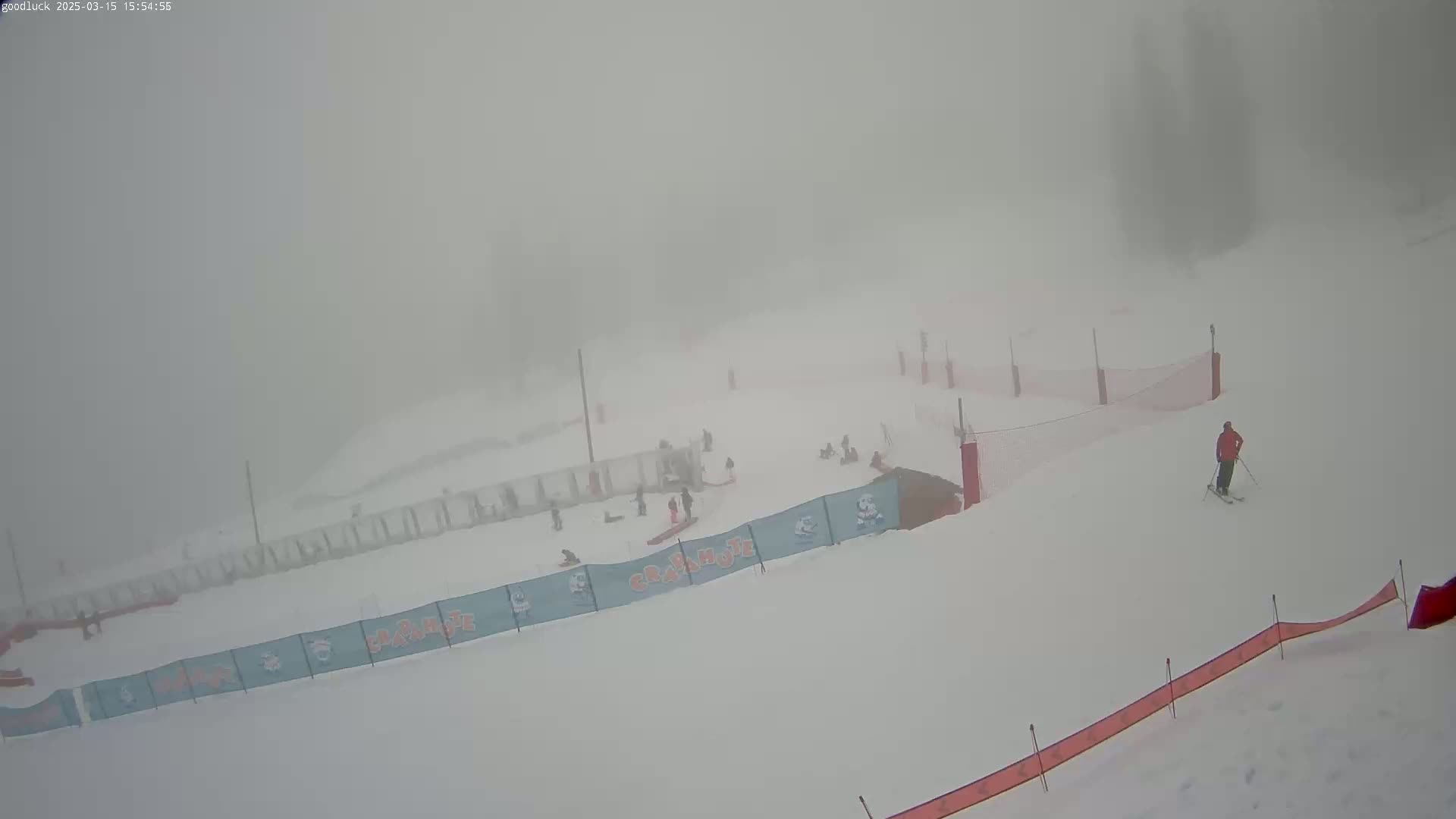 A snowy ski slope is visible through heavy fog, with several skiers scattered across the area.