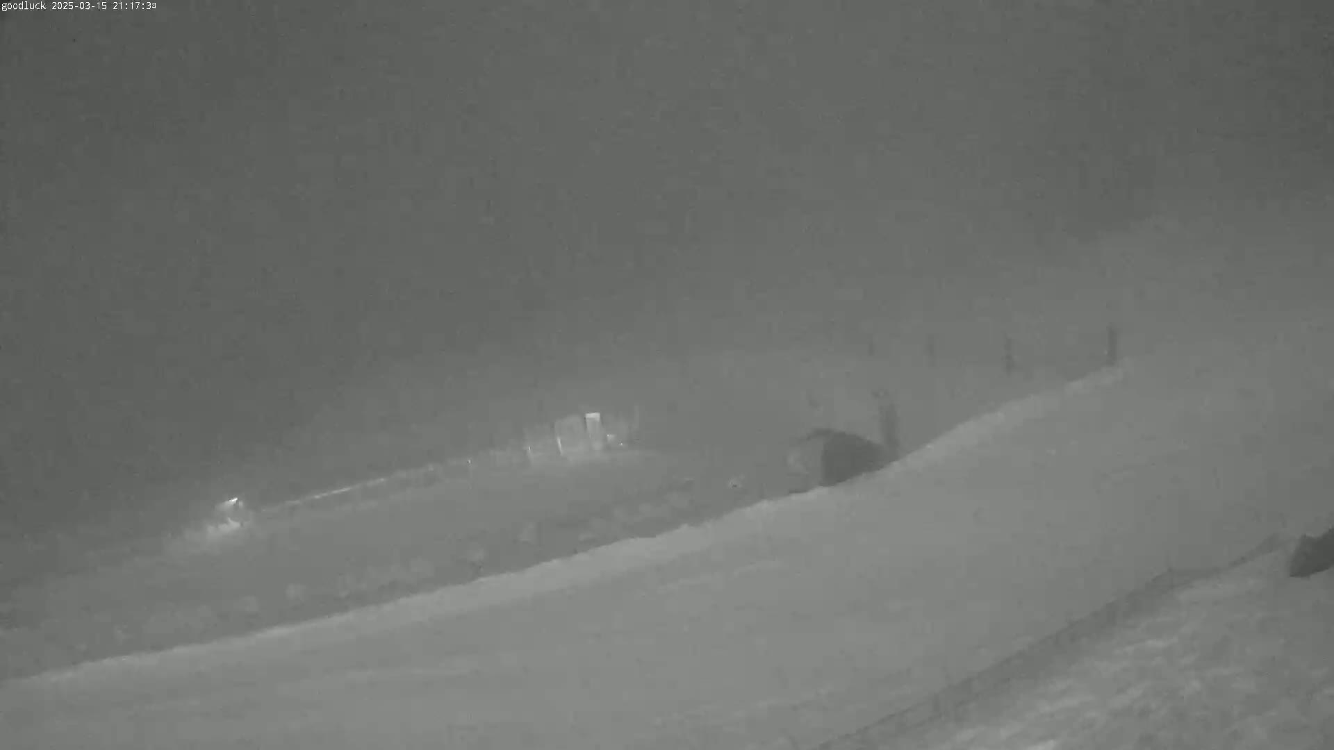 A nighttime, snowy scene shows a sloped, snow-covered area with indistinct structures and figures in the distance under a hazy sky.