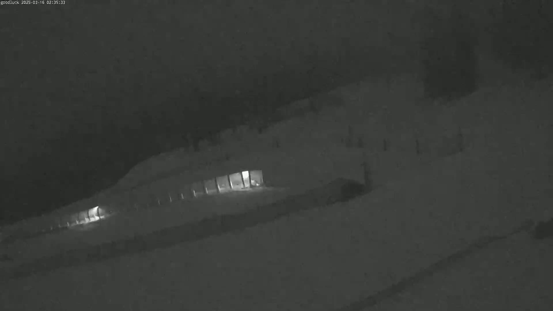 A dimly lit, snow-covered area at night shows a long, low building with lights on inside.