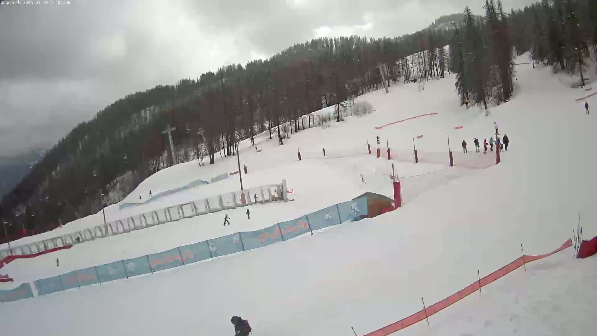 A snowy ski slope with several people skiing and walking, surrounded by a wooded hillside under an overcast sky.