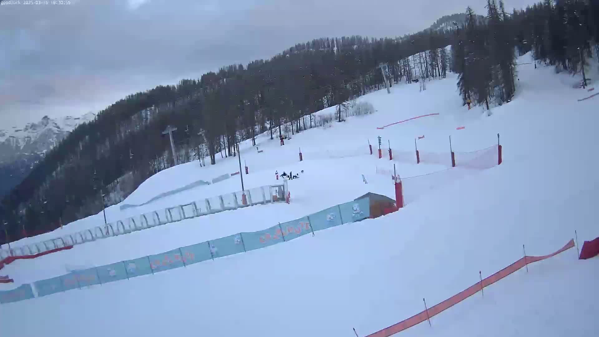 A snow-covered ski slope with several skiers, safety netting, and a few buildings is visible under an overcast sky.