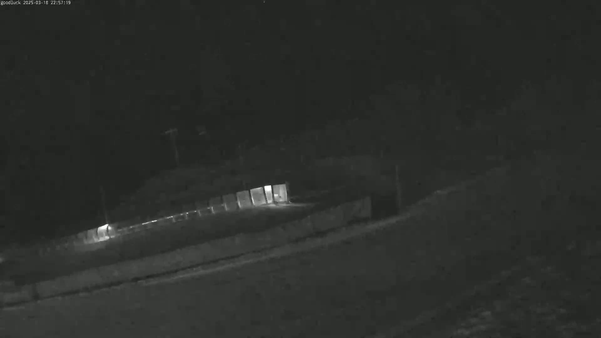 A nighttime, low-light image shows a long, low building with illuminated windows, partially obscured by a fence or wall.