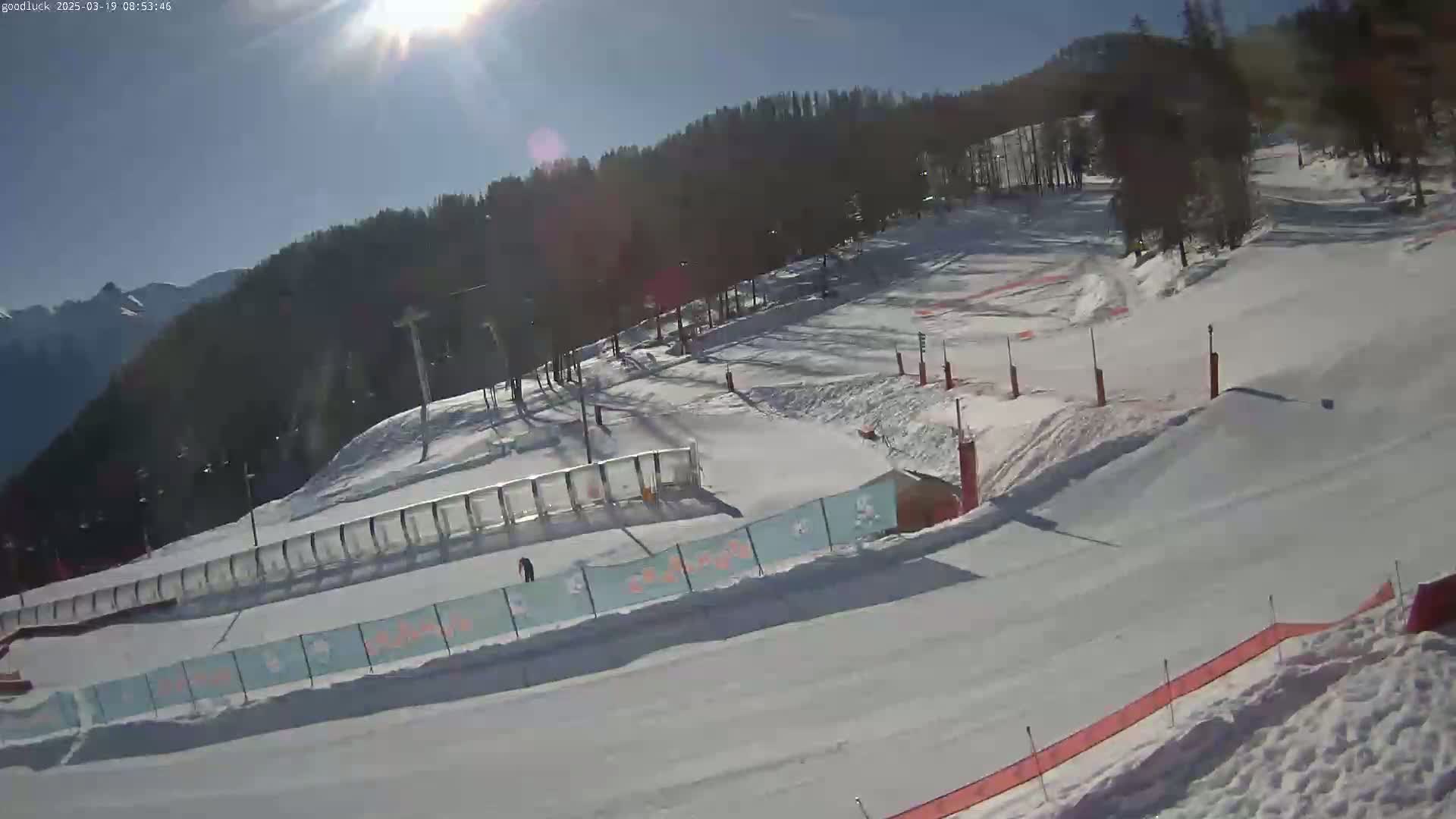 A sunny day at a snow-covered ski slope with a few people visible, featuring ski lifts, fences, and marked runs.