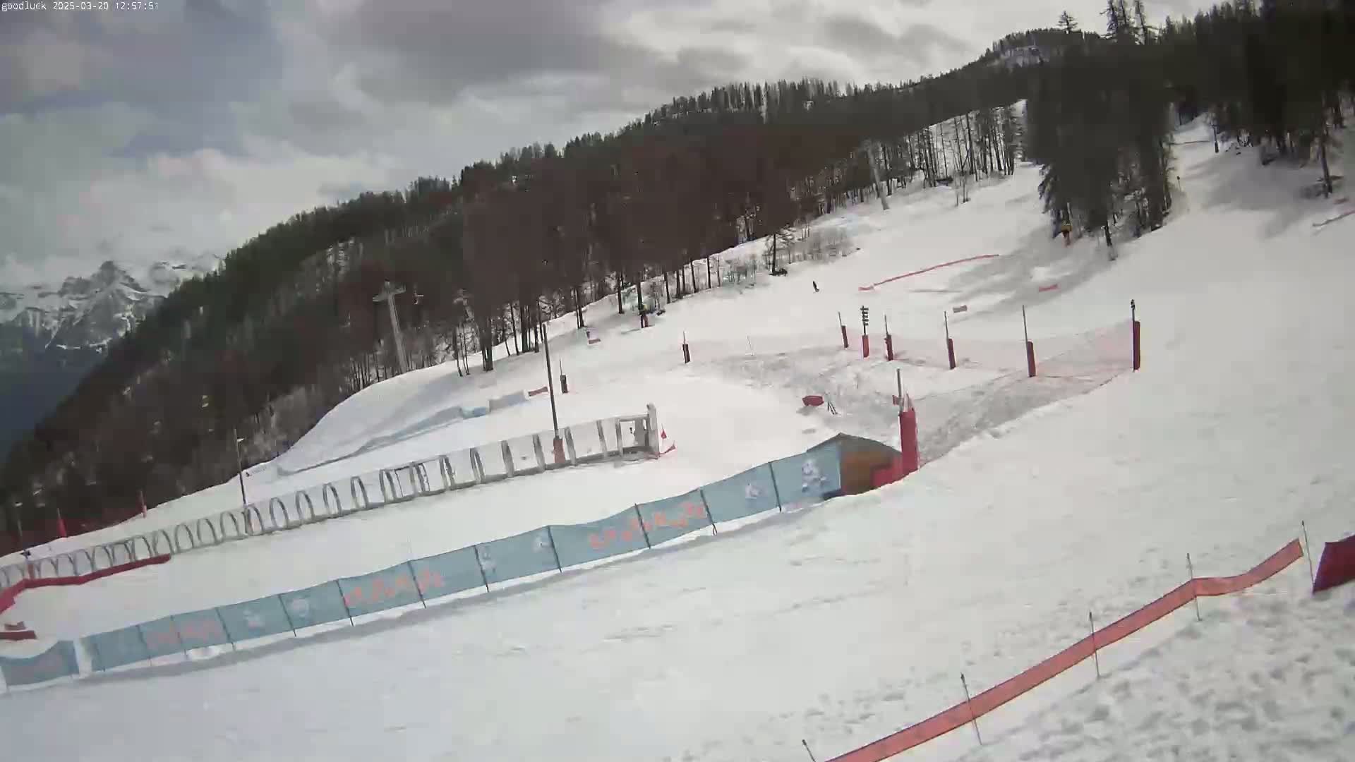 A snow-covered ski slope with a wooded hillside in the background under an overcast sky.