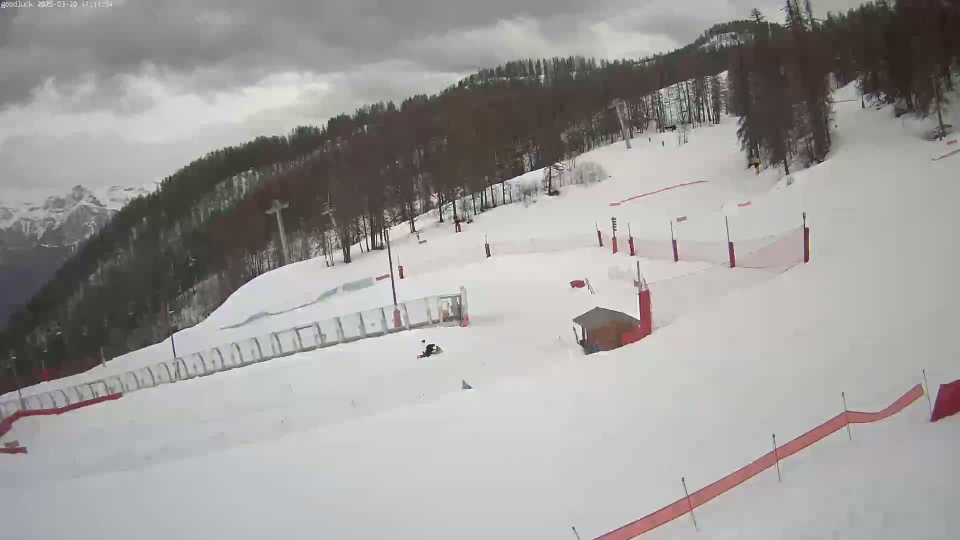 A snow-covered ski slope with several red safety nets and a small building is visible under an overcast sky.