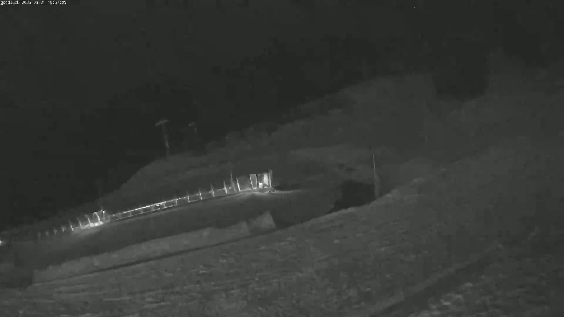 A nighttime, low-light image shows a snow-covered landscape with a fence and some structures in the distance.