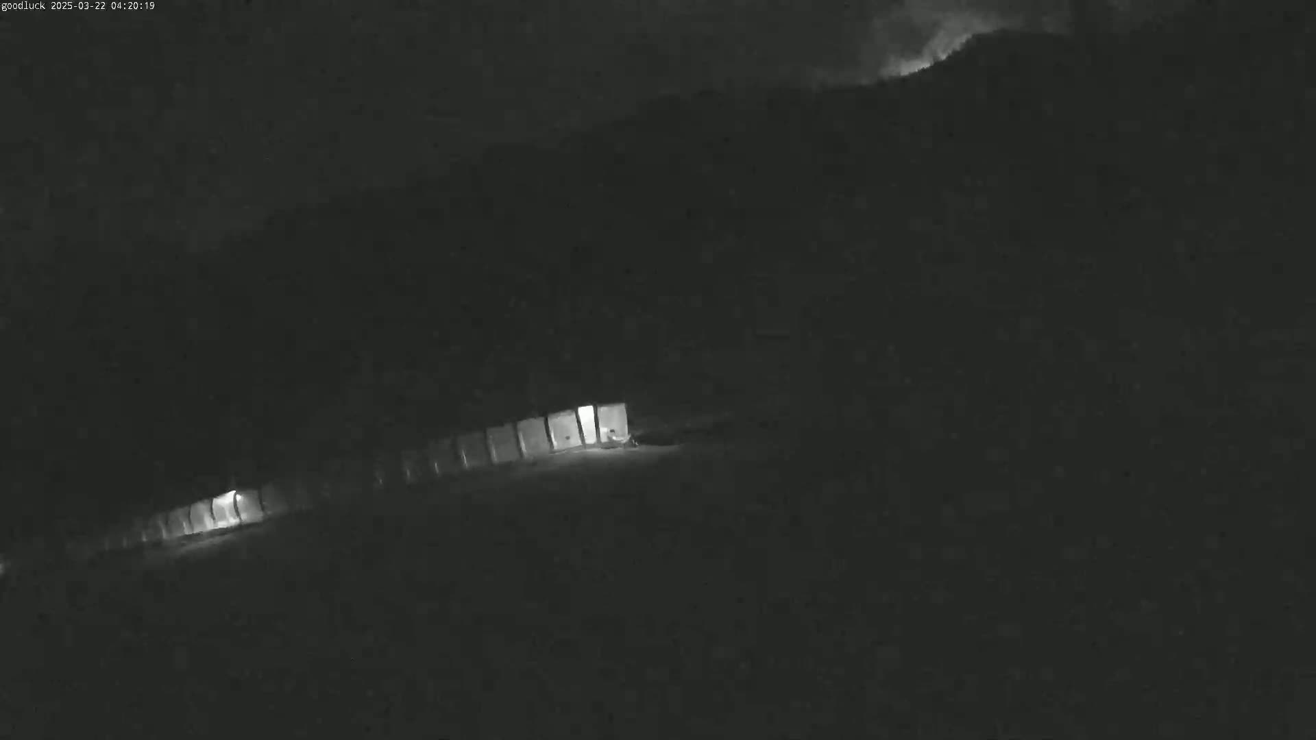 A low-light nighttime image shows a line of illuminated structures at the base of a dark hill under a cloudy sky.