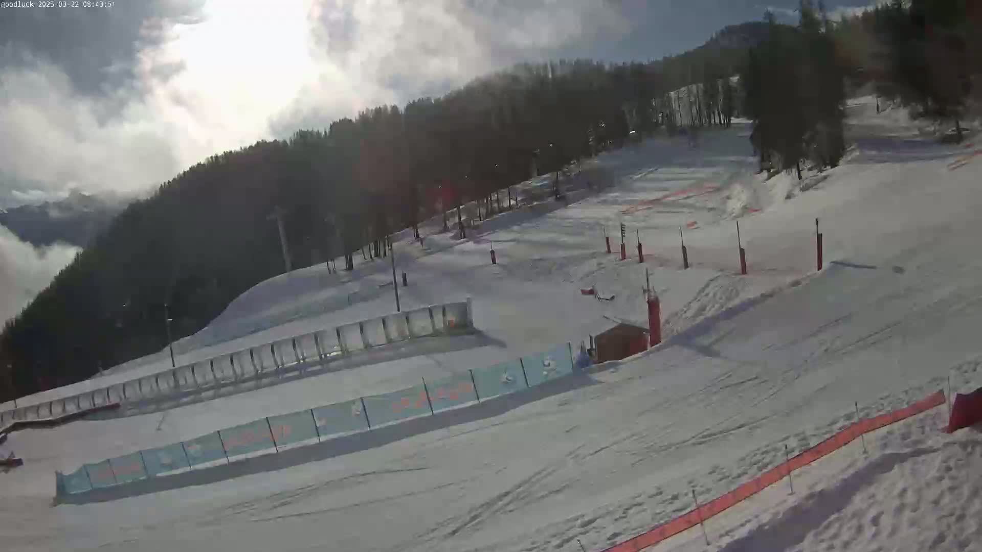 A snow-covered ski slope with trees in the background under a partly cloudy sky.