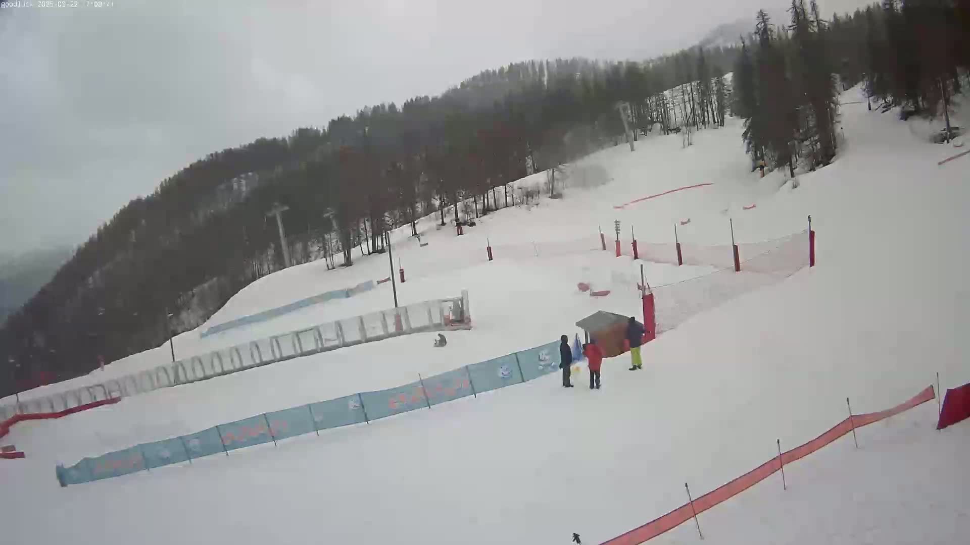 A snow-covered ski slope with several people near safety netting, under an overcast sky.