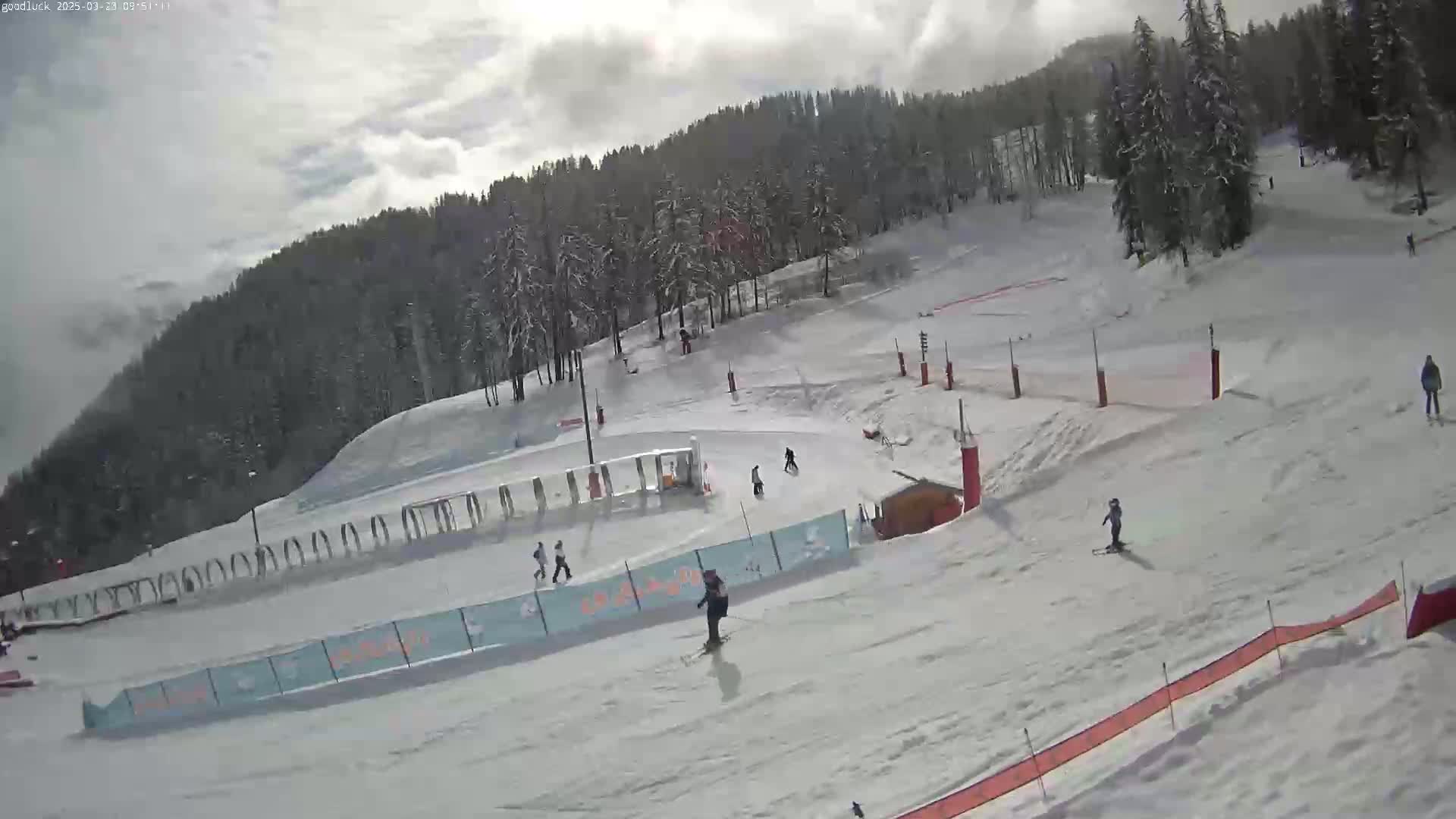 A snowy ski slope with several skiers and snowboarders under a partly cloudy sky.