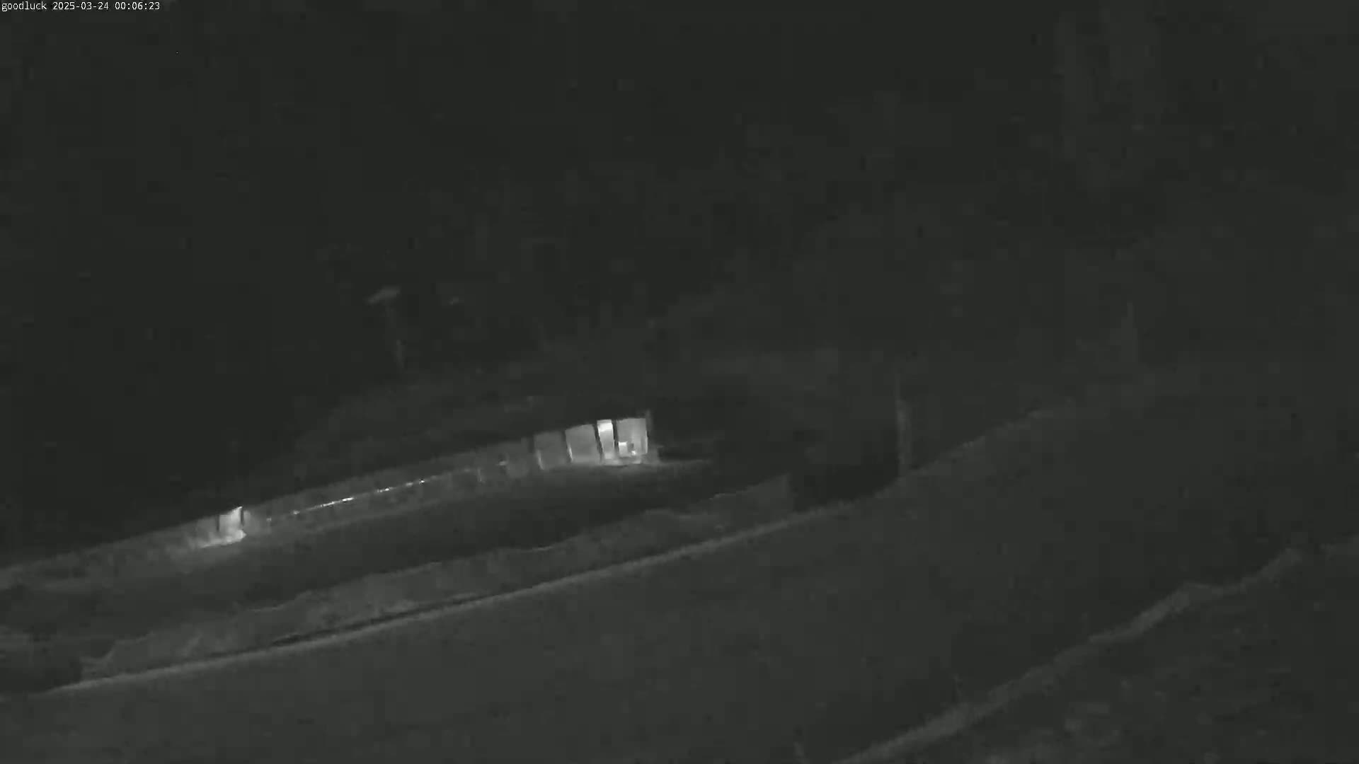A low-light nighttime image shows a dimly lit building and a road, with some vegetation visible.