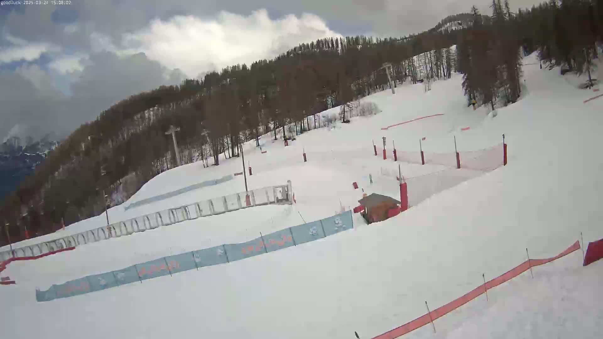 A snow-covered ski slope with trees in the background under a partly cloudy sky.