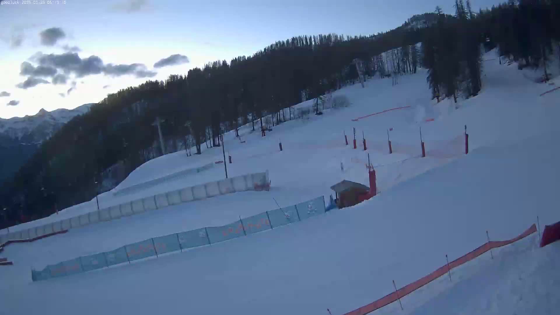A snow-covered ski slope with a few structures and fences is visible under a partly cloudy sky at dawn.