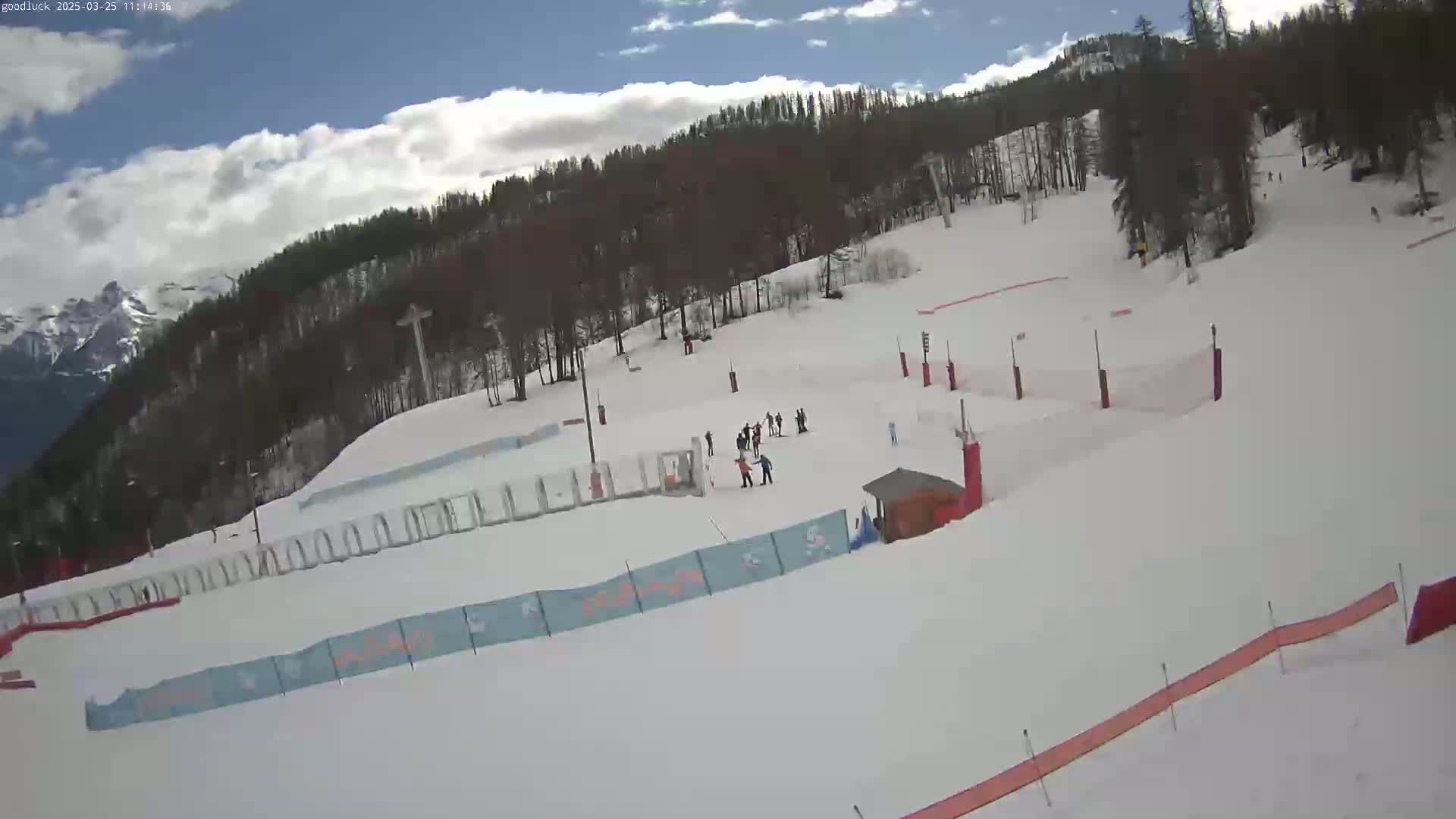 A snow-covered ski slope with several skiers, a small building, and trees in the background under a partly cloudy sky.