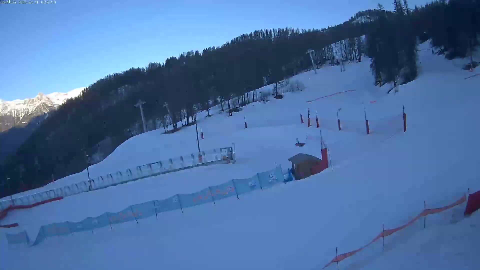 A snow-covered ski slope with a small building and fencing, backed by a dark evergreen forest under a clear blue sky.