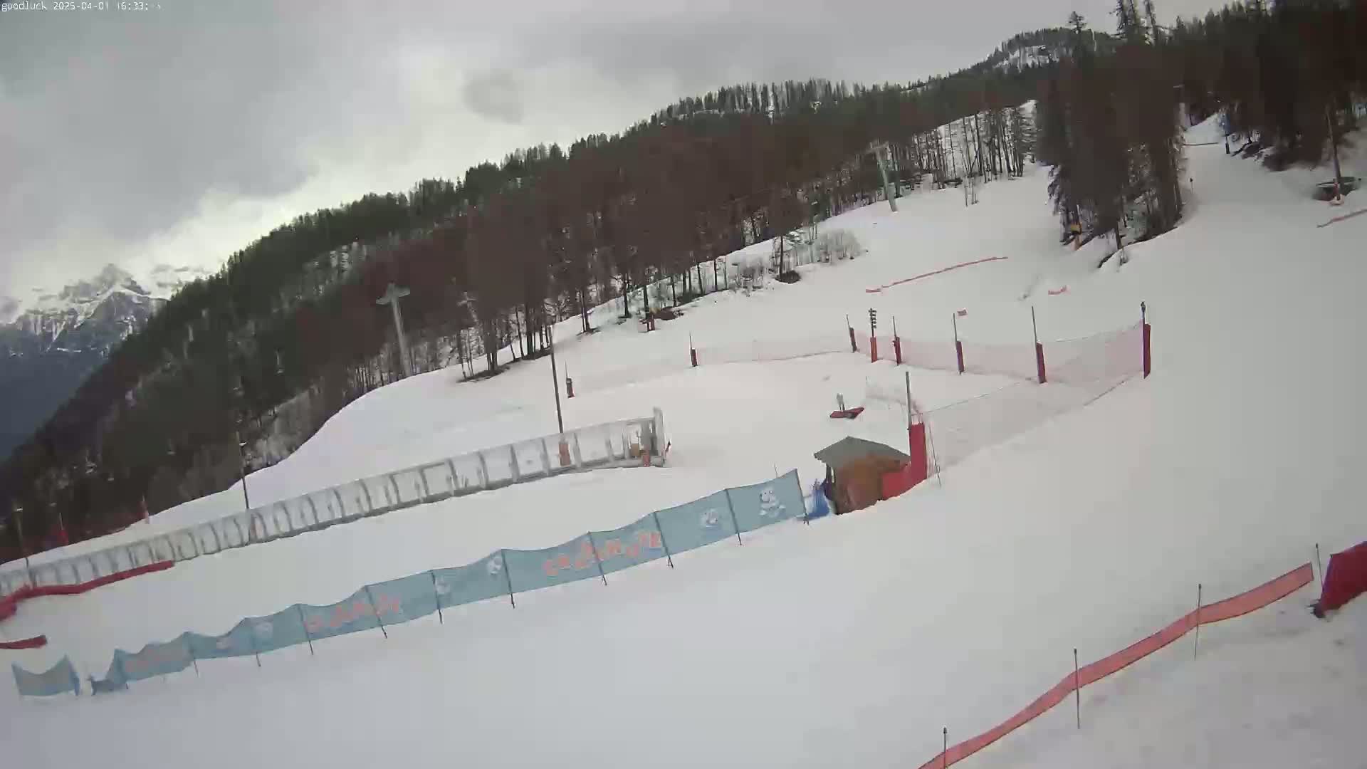 A snow-covered ski slope with several fences and a small building is visible under an overcast sky.