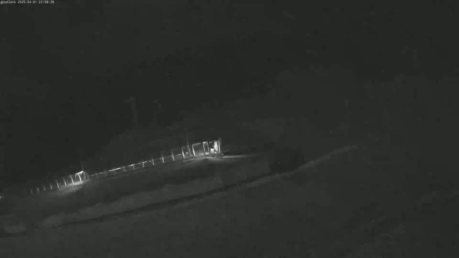 A low-light, nighttime image shows a building with a fence and some snow or ice on the ground.
