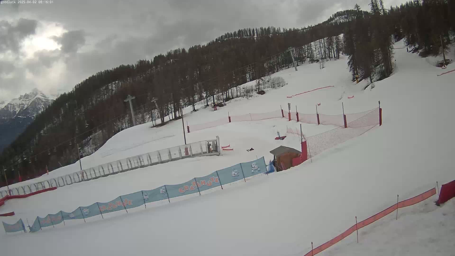 A snow-covered ski slope with a few people, safety netting, and a small building under an overcast sky.