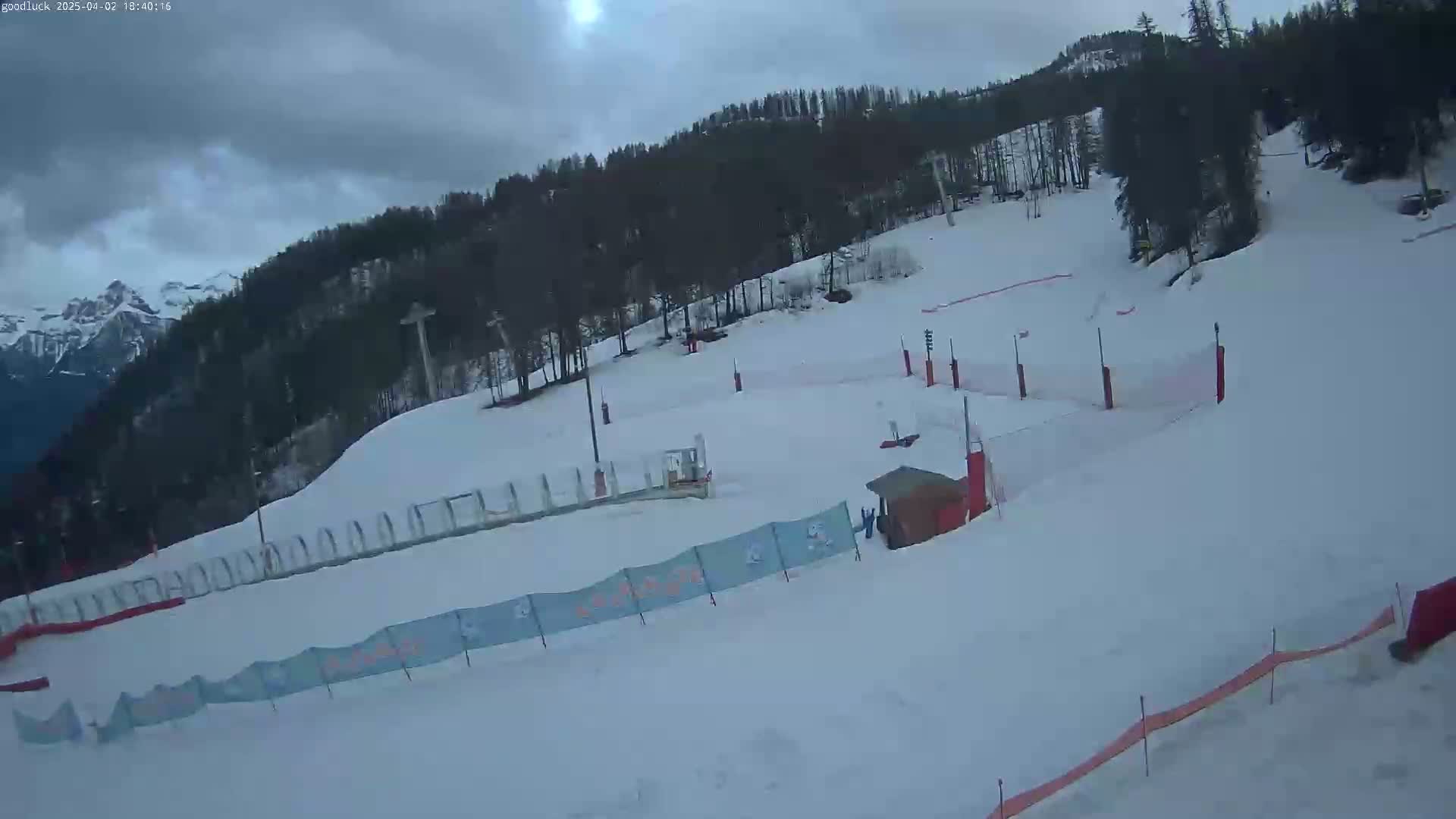 A snowy ski slope with a small building and safety nets is visible under an overcast sky.