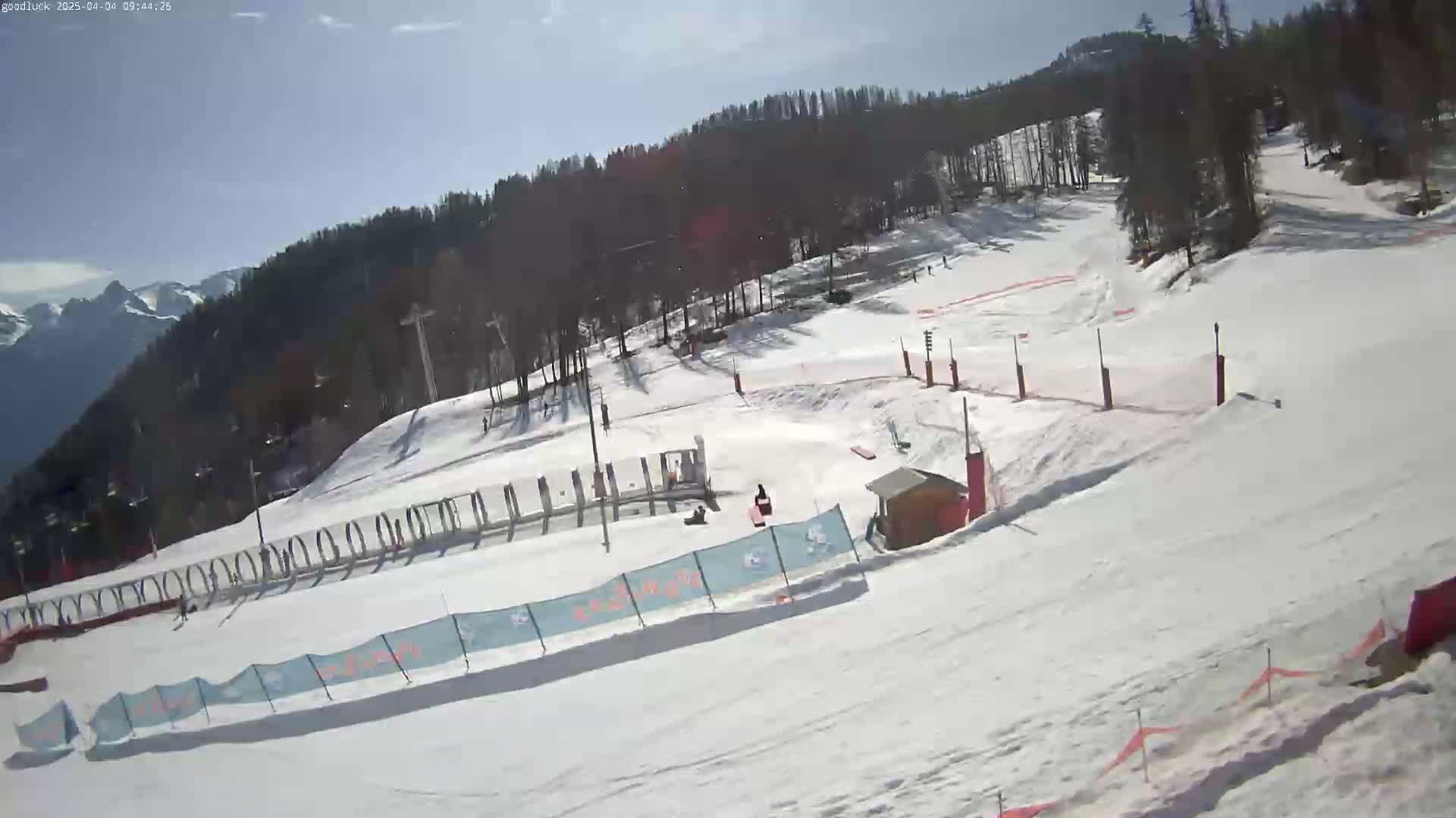 A snowy ski slope with a few people visible on a sunny day, surrounded by a treeline and mountains in the distance.
