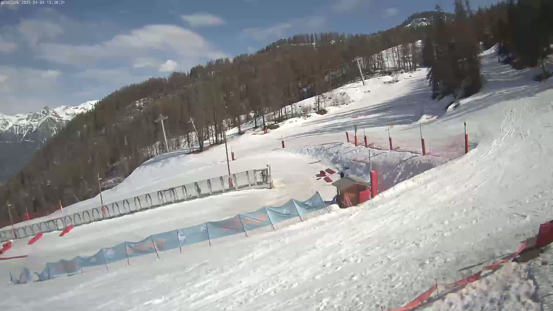 A snow-covered ski slope with sparsely populated trees in the background under a partly cloudy sky.