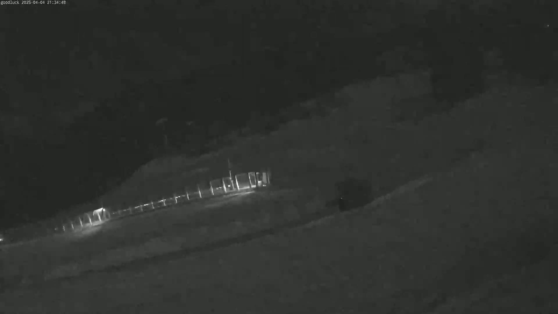 A nighttime, low-light image shows a dimly lit structure and fence near a snow-covered expanse.