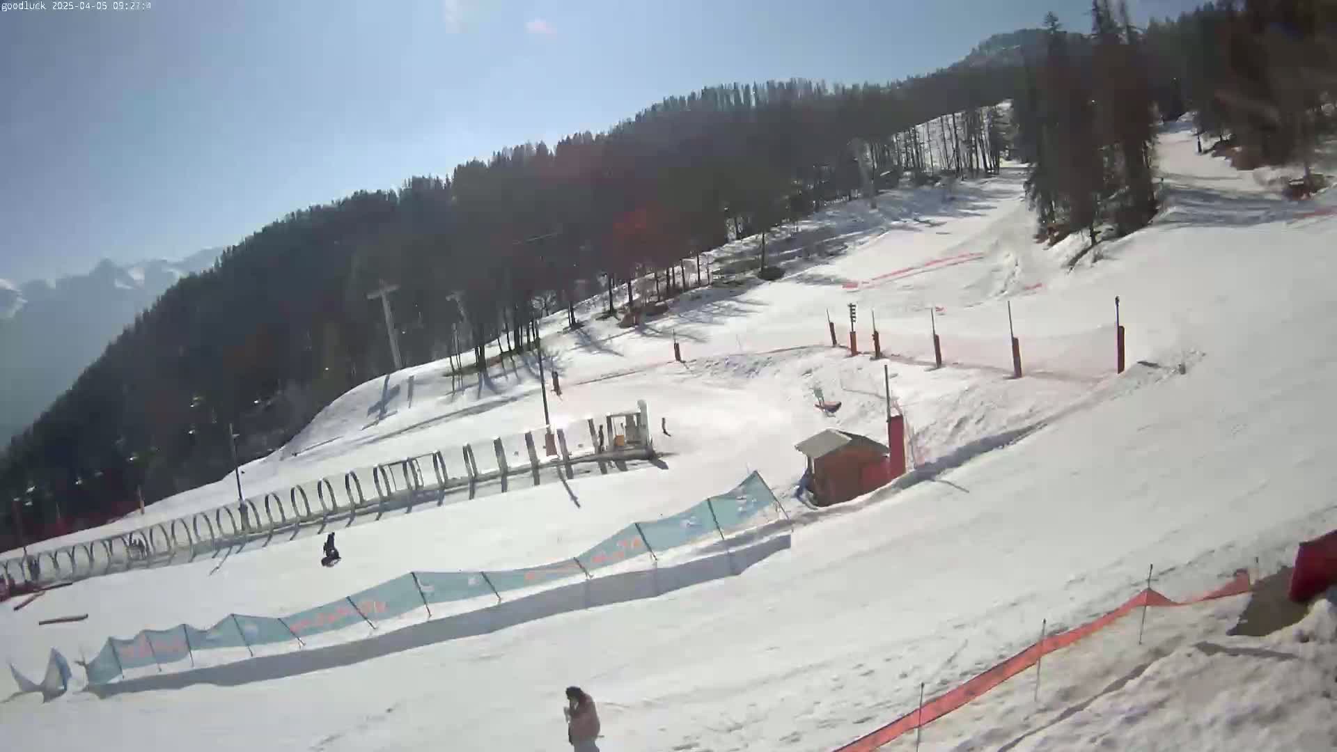 A snowy ski slope on a sunny day, with several skiers visible and a lift in the background.