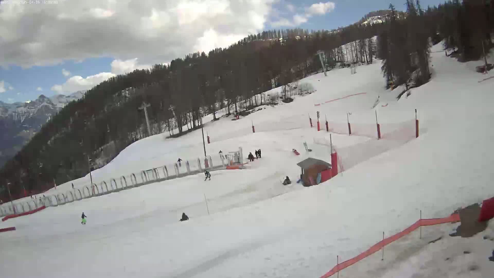 A mostly sunny day reveals a snow-covered ski slope with several people skiing and snowboarding, surrounded by trees and mountains in the background.