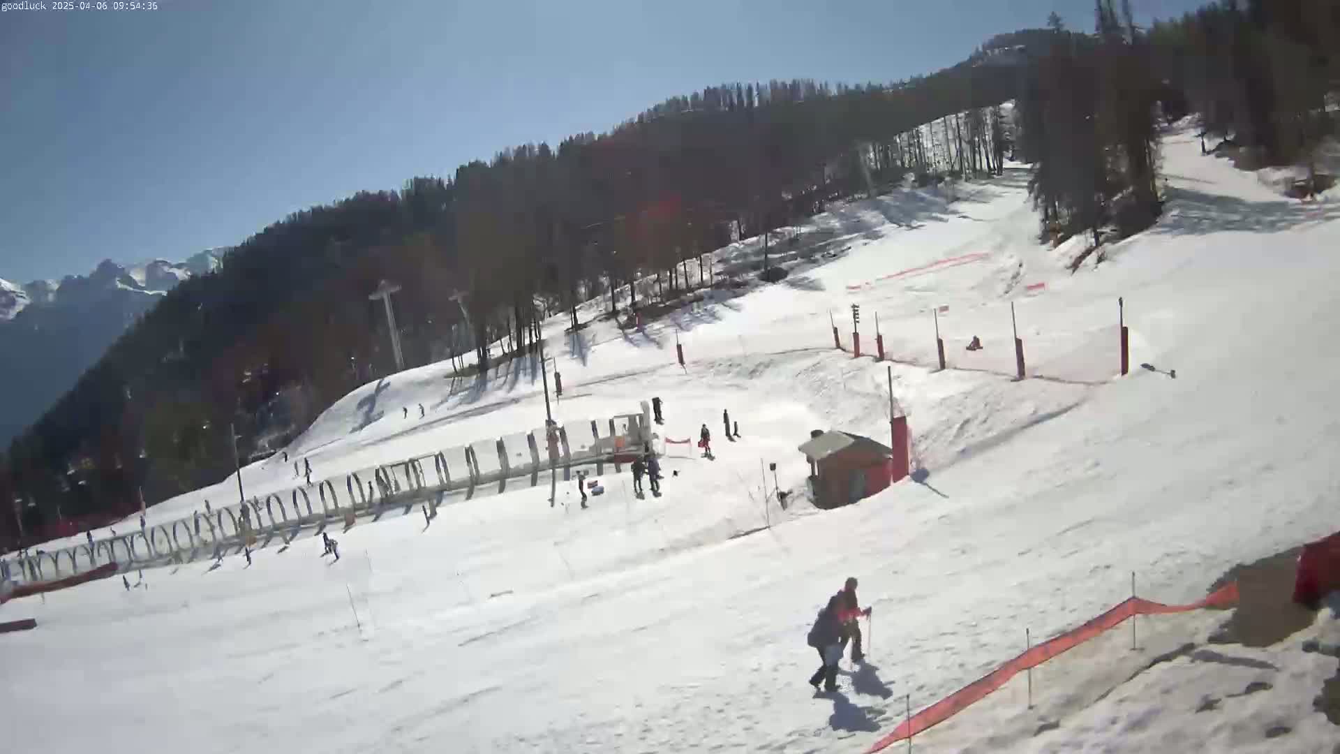 A sunny day at a snow-covered ski resort shows several people skiing and snowboarding down a slope, with ski lifts and other infrastructure visible.