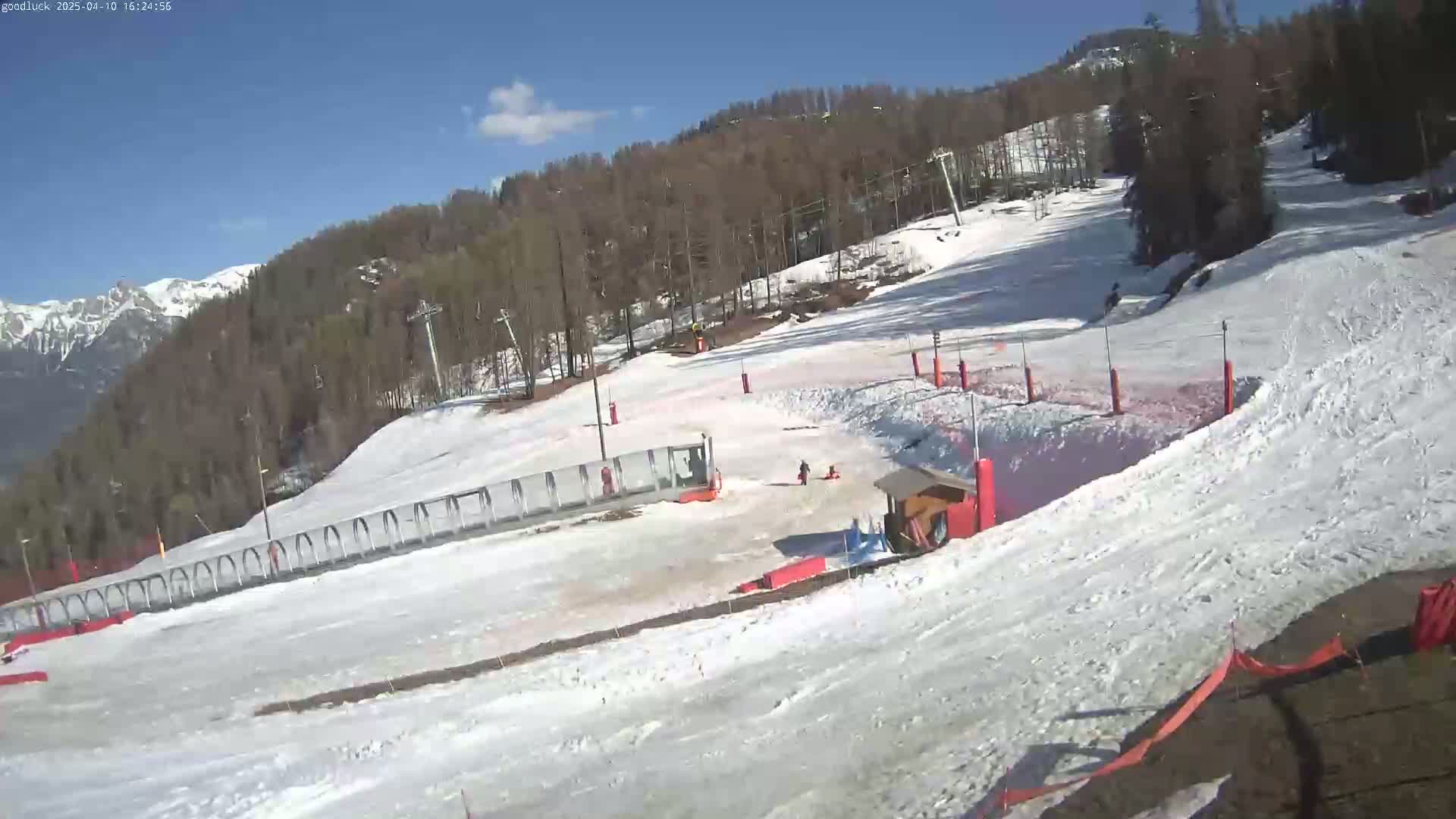 A snow-covered ski slope with several people and equipment is visible under a bright, sunny sky, with mountains in the background.